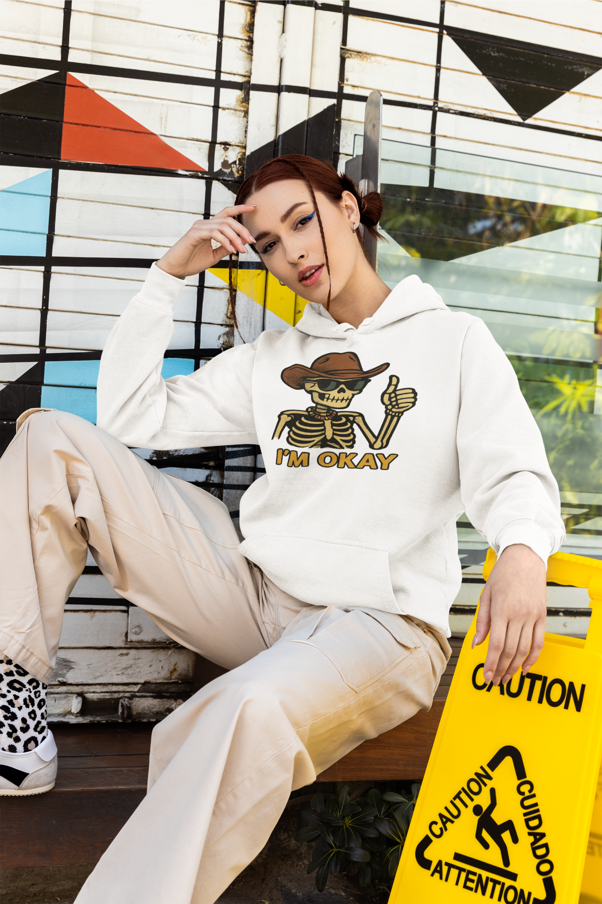 Mashafrica white hoodie with skeleton cowboy graphic and Im Okay text model seated casually outdoors